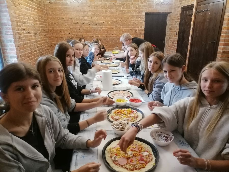 Uczniowie siedzący i przygotowują pizze przy długim stole.